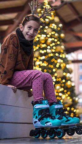 Comprar patines para niños - Niña sentada sonriendo con patines puestos y un árbol de navidad de fondo